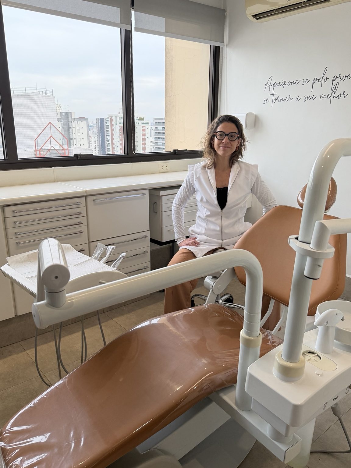 Dra. Camila Meira sorrindo, sentada em seu consultório odontológico com vista para a cidade de São Paulo