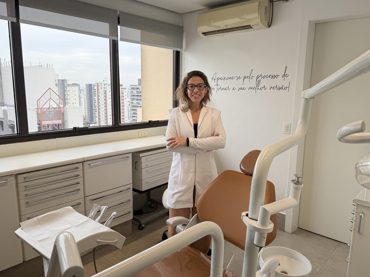Dra. Camila Meira em pé no consultório, ao lado da cadeira odontológica